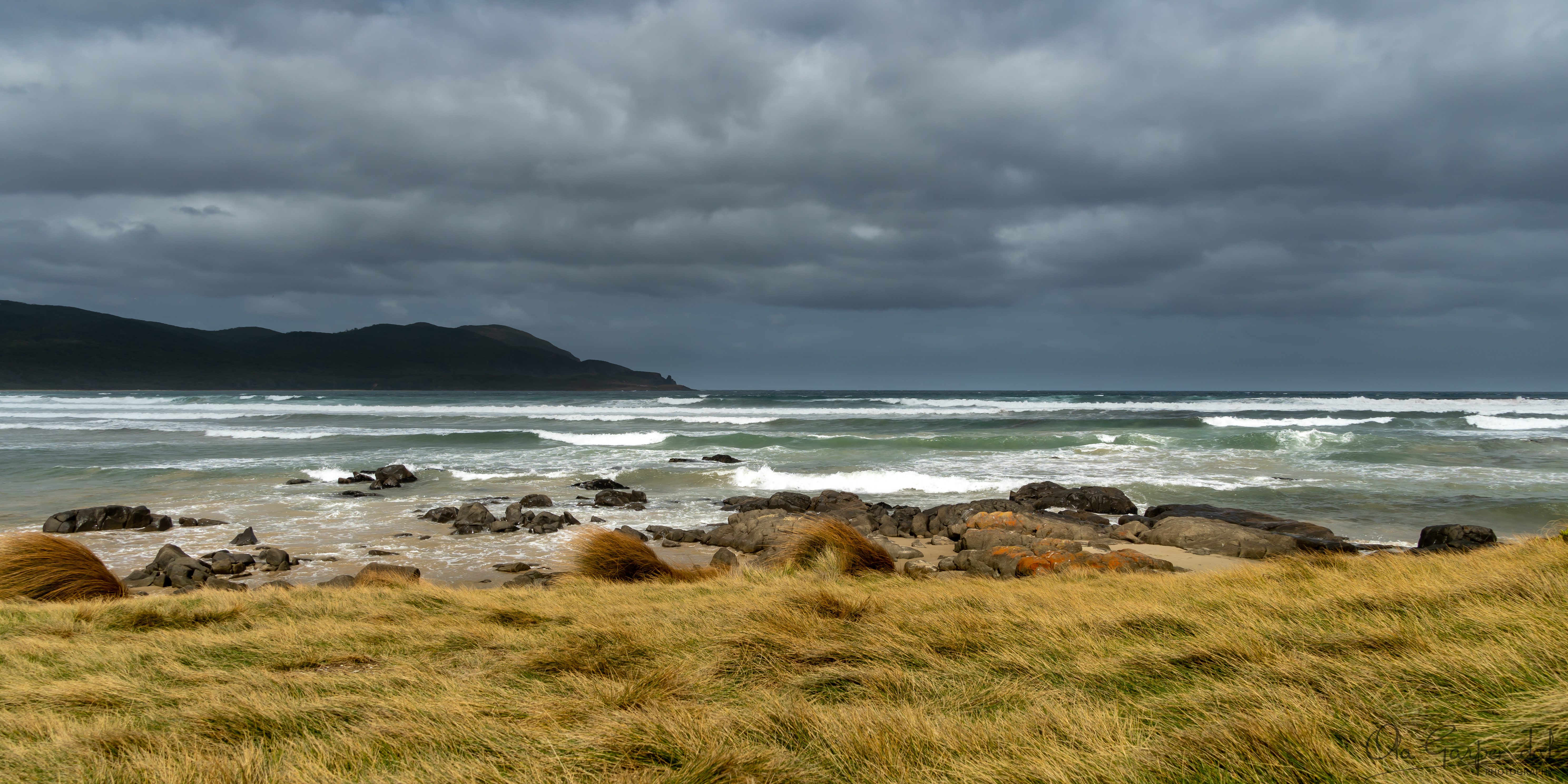 Storm at Cloudy Bay showing wind-swept coastal grasses, rocky shoreline, rolling surf, distant headlands and dramatic storm clouds over southern Bruny Island in March, photographed by Ola Garpendal Photography