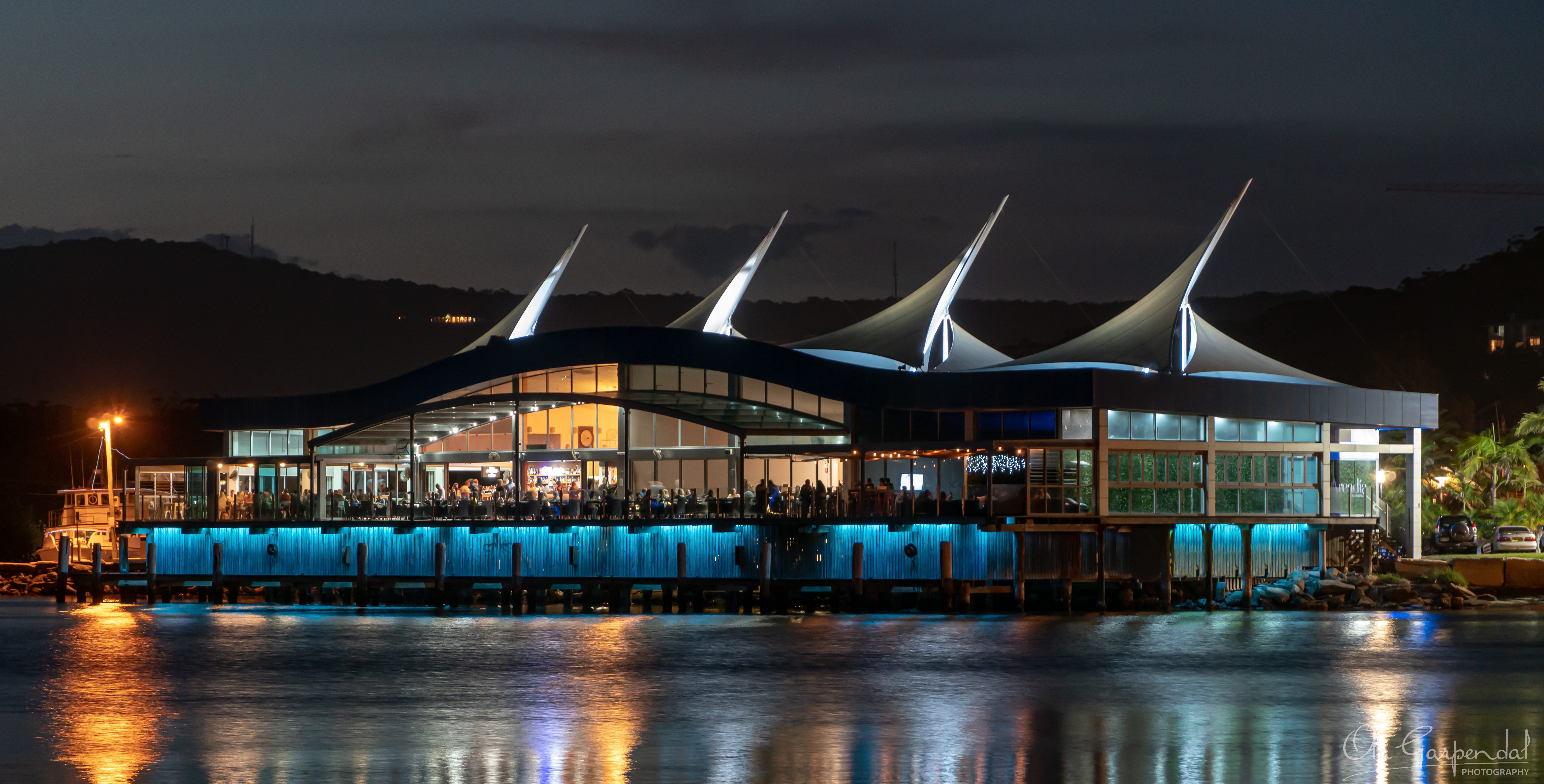 Gosford Waterfront at Night showing the illuminated sail-roof waterfront restaurant at Gosford, with warm interior lighting, diners, colourful reflections on calm water and a dark spring evening sky, photographed by Ola Garpendal Photography