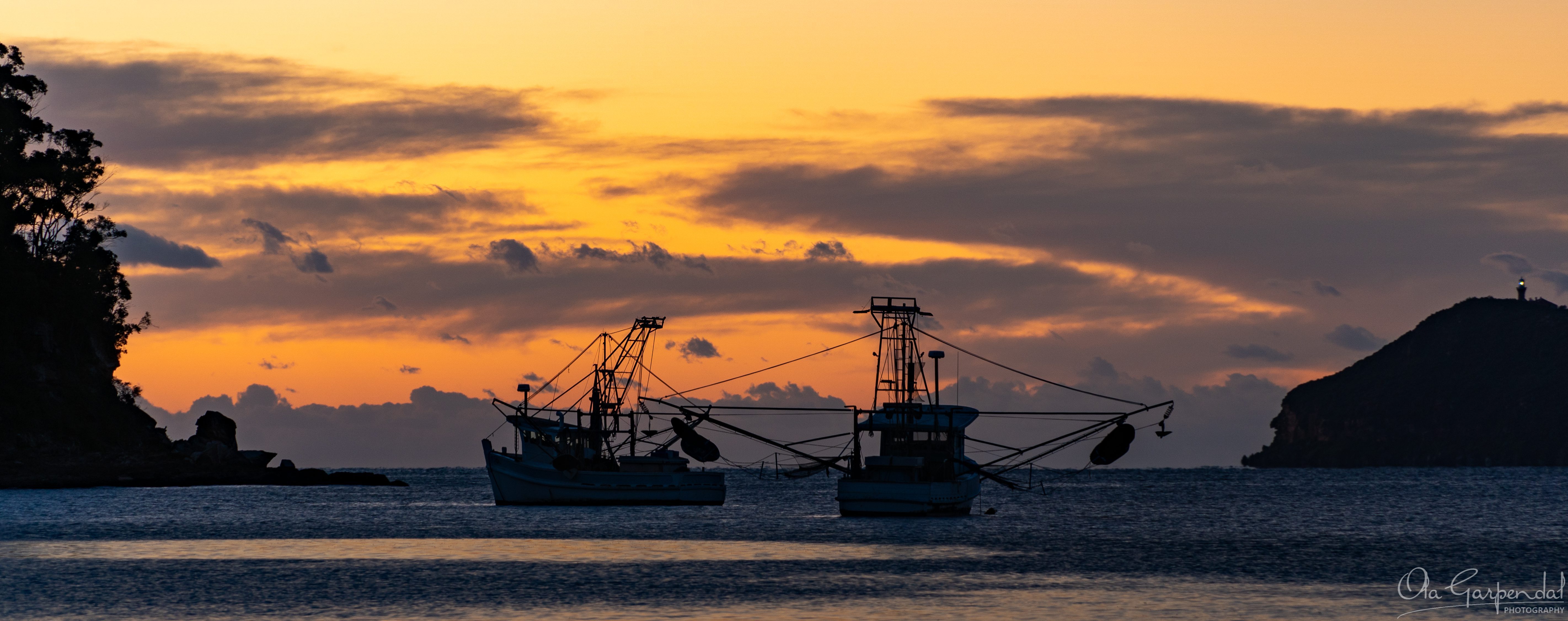Fishing Boats at Patonga shows two fishing boats silhouetted on calm water before sunrise, with warm dawn colours, Broken Bay, the Hawkesbury River entrance and distant Barrenjoey Lighthouse, photographed by Ola Garpendal Photography