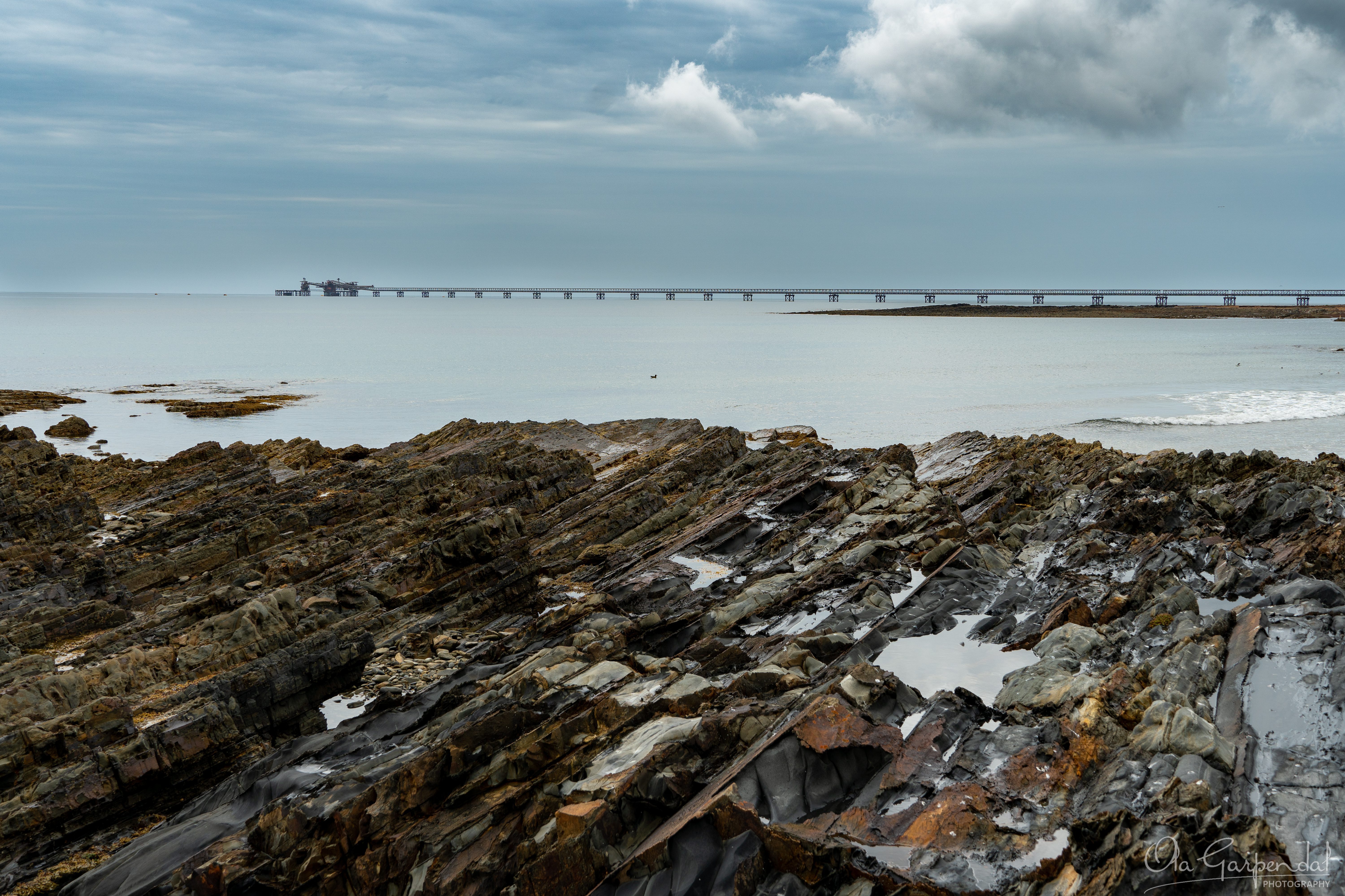 Port Latta Jetty showing layered rocky shoreline in northern Tasmania, calm coastal waters, a long industrial jetty extending into Bass Strait, and dramatic cloud-filled skies in March, photographed by Ola Garpendal Photography