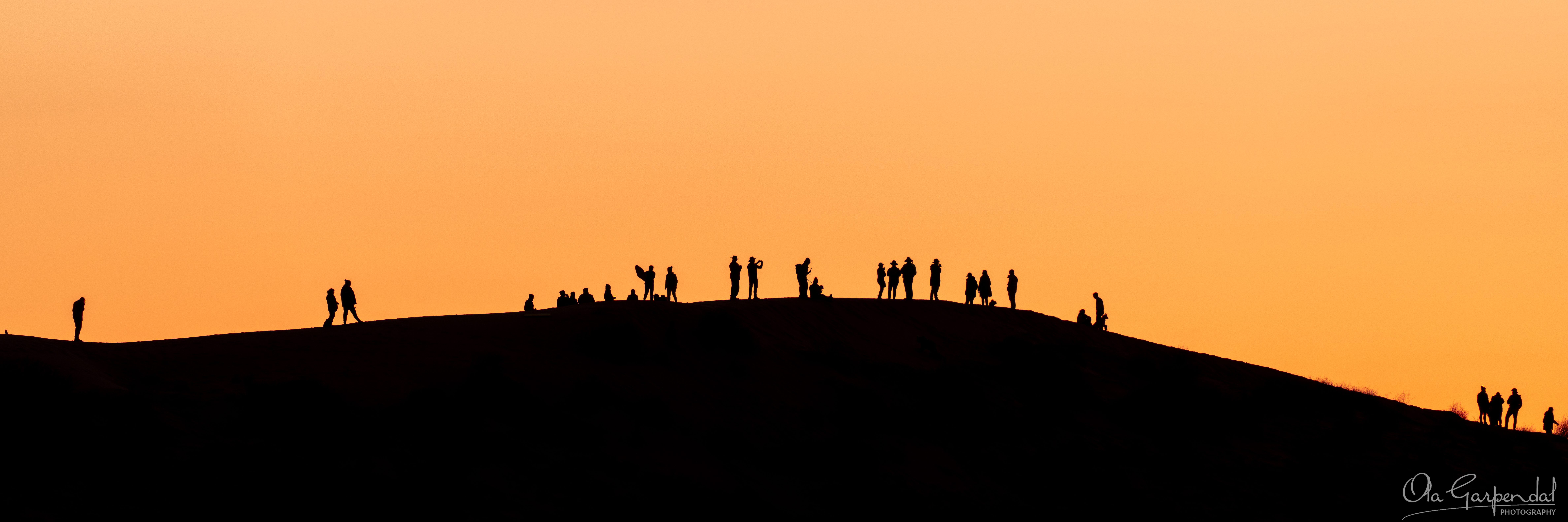 Big Red Silhouette showing visitors standing along the crest of Big Red near Birdsville at sunset, with dark dune silhouettes against a glowing orange July evening sky, photographed by Ola Garpendal Photography