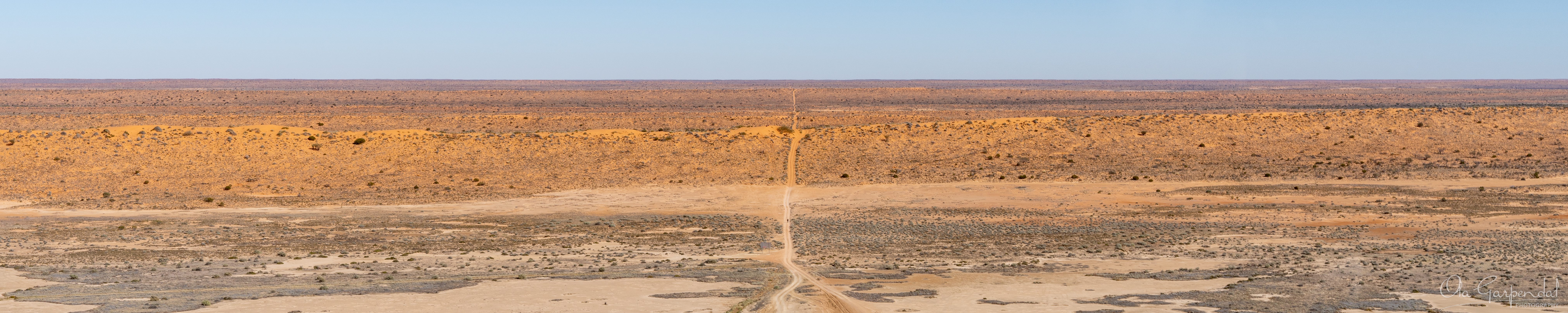 Road to Simpson Desert aerial panorama near Big Red and Birdsville showing a remote desert track crossing dunes and arid plains beneath a distant horizon in July, photographed by Ola Garpendal Photography