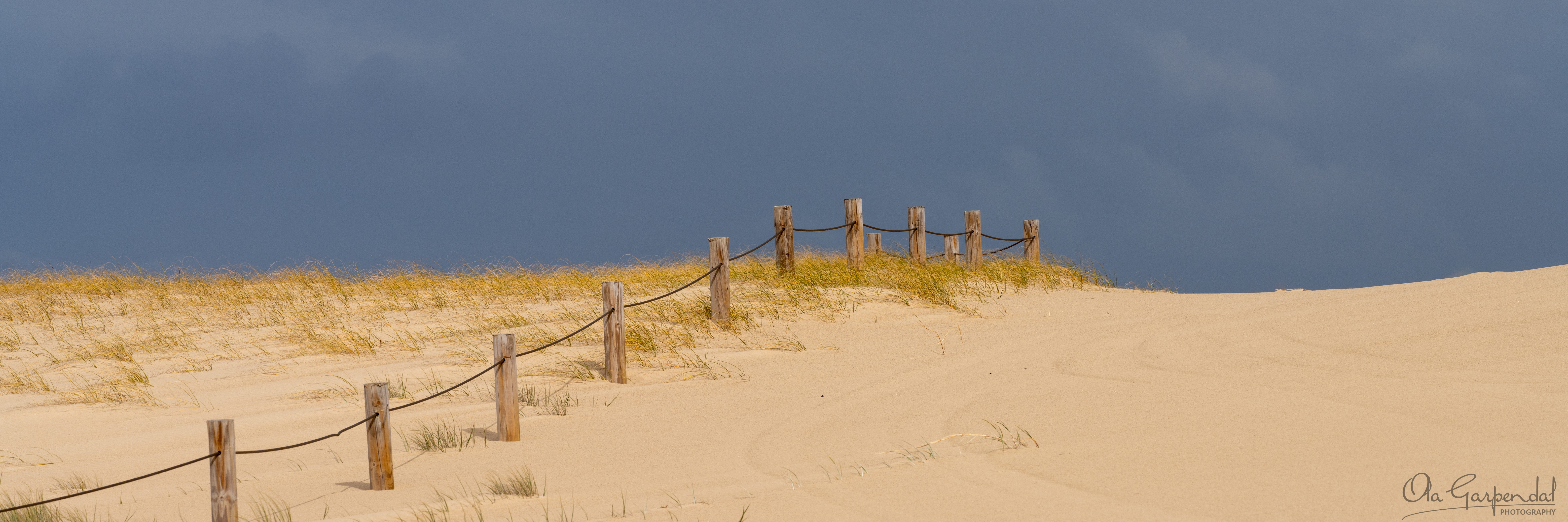 Off Limits! — a weathered rope and timber fence climbing a coastal sand dune toward the horizon, with golden grass and a storm-darkened sky, photographed by Ola Garpendal Photography.
