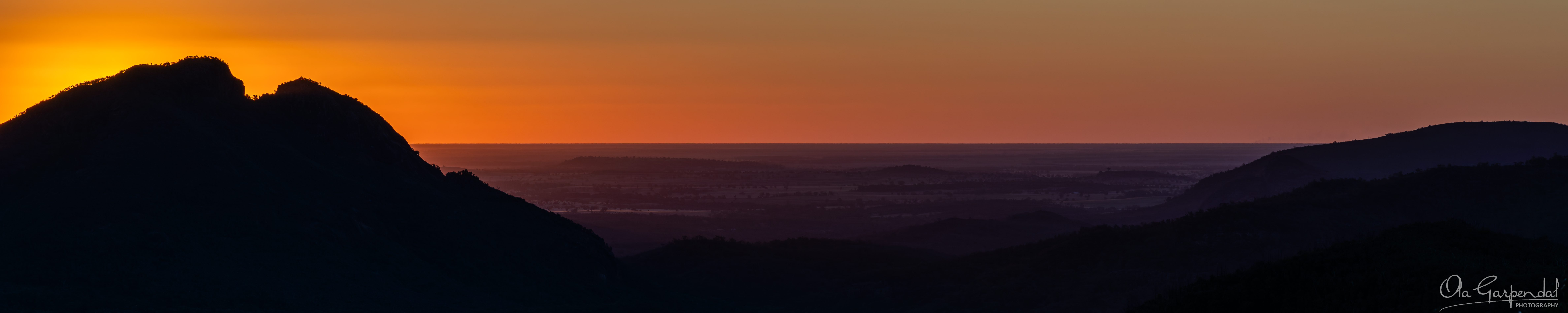 Split Rock Sunset — the silhouette of Split Rock in Warrumbungle National Park against a deep orange and amber sunset sky, with layered purple ridgelines receding across the central-western NSW plains, photographed by Ola Garpendal Photography.