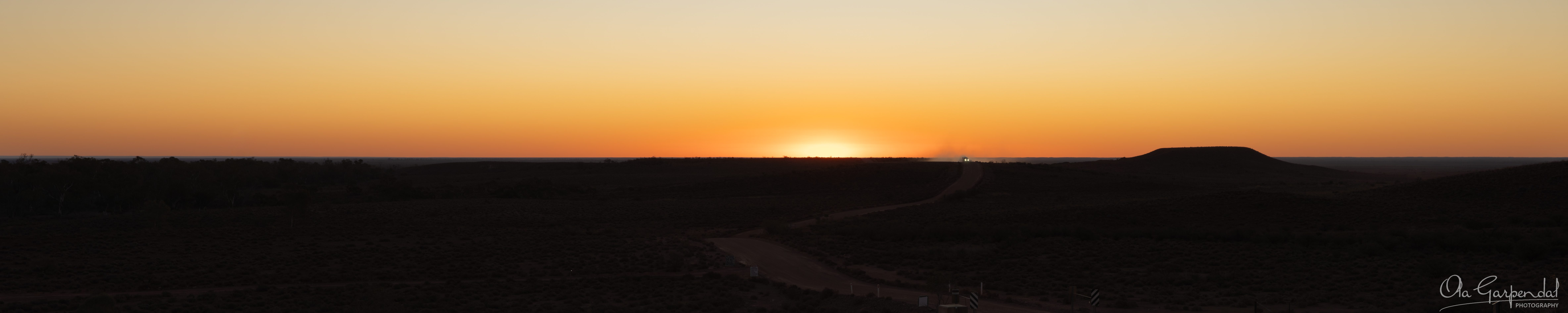 Late Arrival showing distant vehicle headlights and a trail of dust on Brachina Gorge Road beneath a glowing amber sunset sky in the Flinders Ranges, with silhouetted mesas and outback landscapes at dusk, photographed by Ola Garpendal Photography