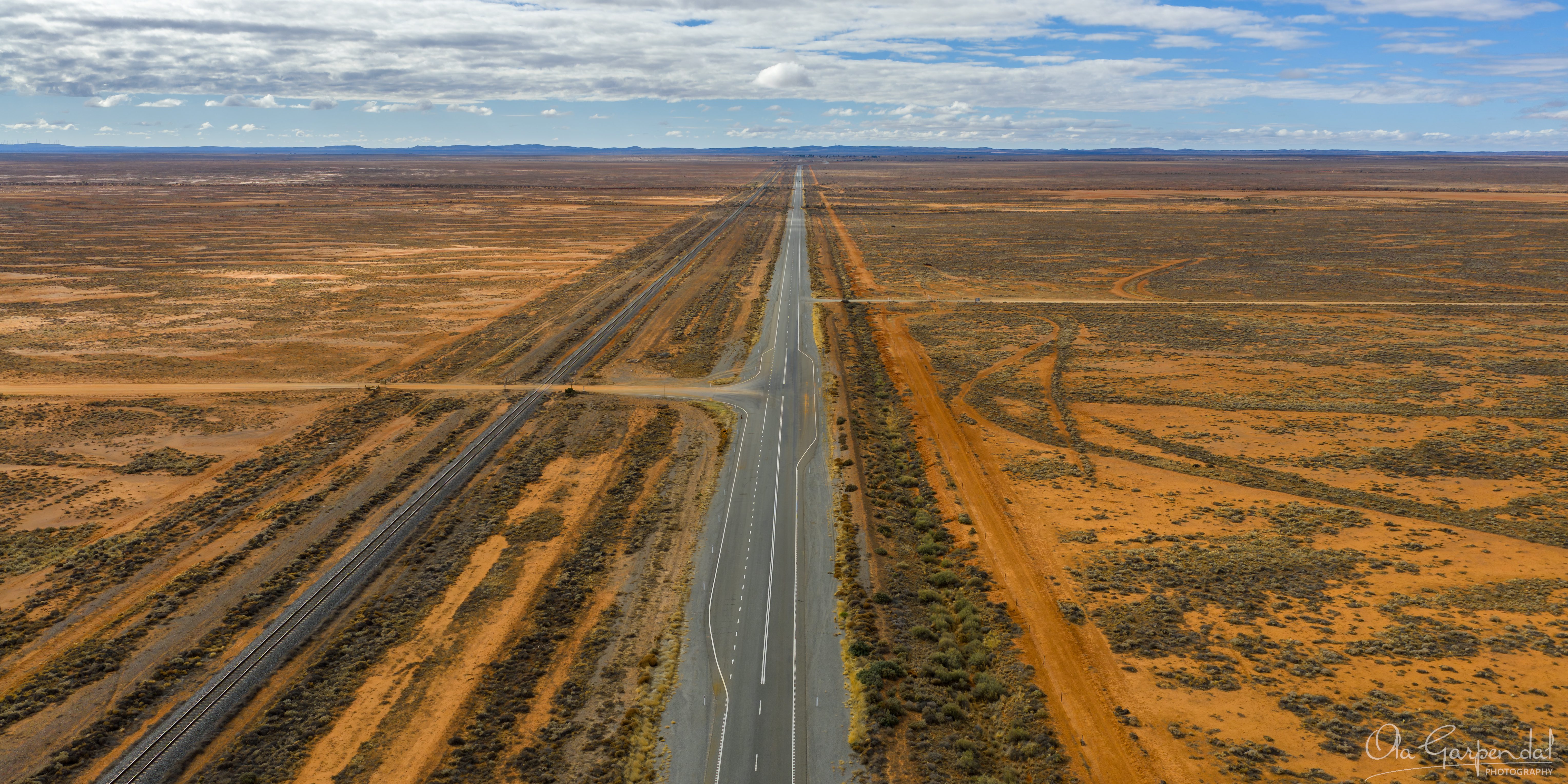 A long, straight road disappearing into the distance. The ground along the road is covered in red dirt. Above the mountain range in the distance is a blue sky with some clouds.