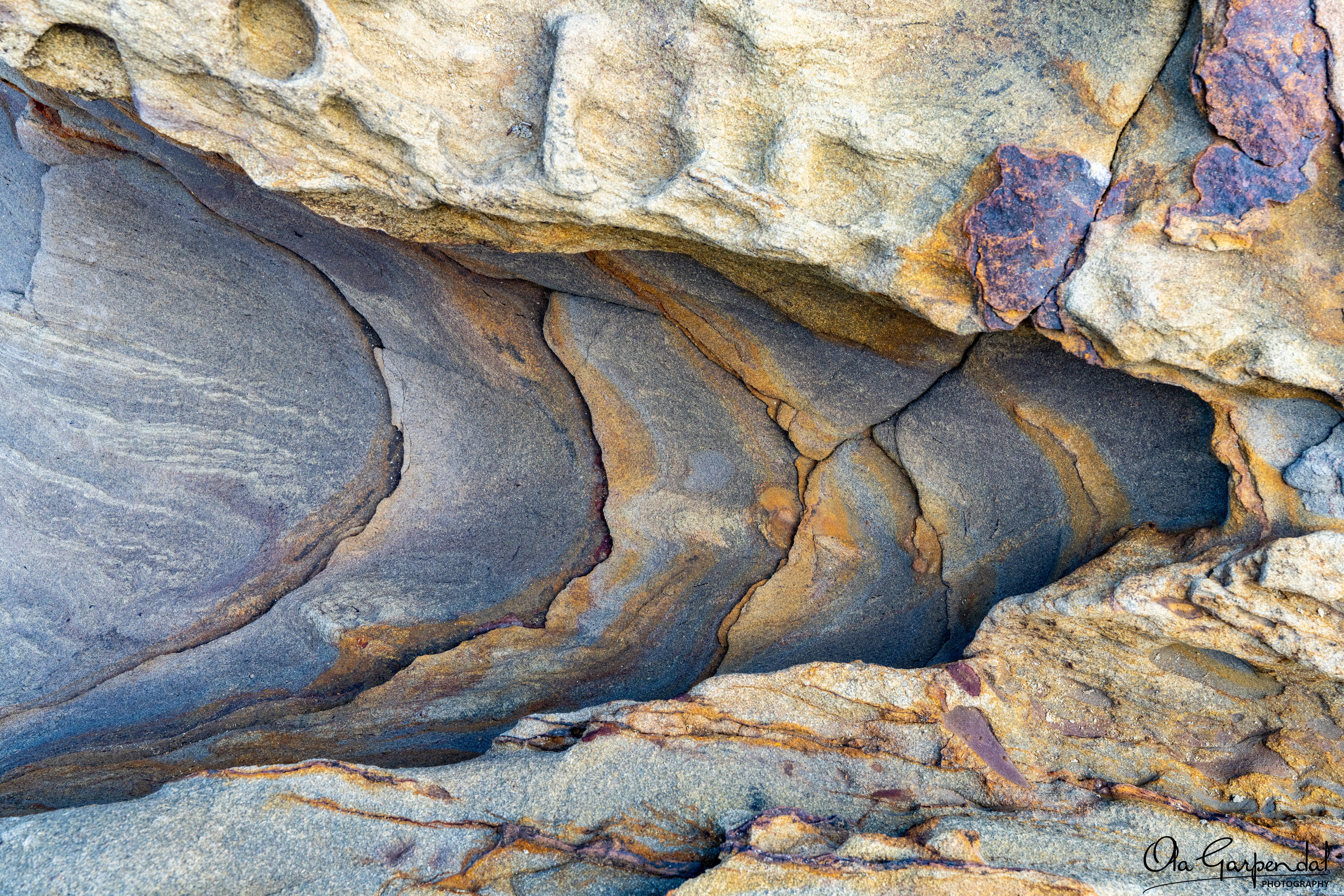 “Dragon Tail” — a close‑up abstract view of curved sedimentary rock layers in grey, tan and rust tones, photographed by Ola Garpendal Photography
